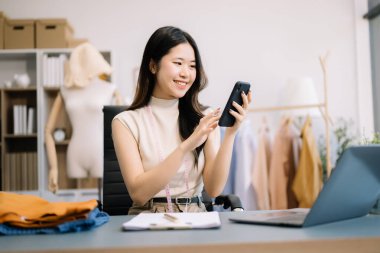 Asyalı terzi kadın terzi atölyesinde giysiler üzerinde çalışıyor. Modern stüdyoda başarılı olduktan sonra güzel genç bayan moda tasarımcısı gülümsemesi