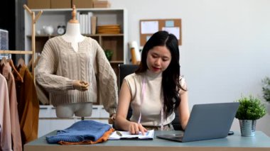  Moda tasarımcısının masasındaki Asyalı genç kadın stüdyoda dizüstü bilgisayarda çalışıyor.