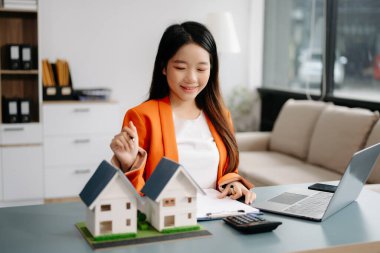 Modern ofiste dizüstü bilgisayarla çalışan genç bir emlakçı ve yanında küçük evler modeli.