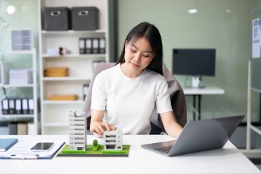 Genç emlakçı ofiste dizüstü bilgisayarla ve yanında küçük ev modeliyle çalışıyor.