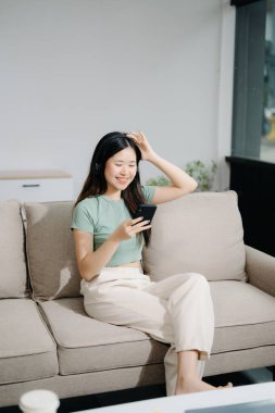 Genç Asyalı kadın evdeki kanepede dinleniyor ve akıllı telefon kullanıyor.