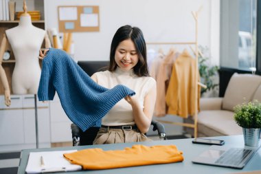 Asyalı terzi kadın terzi atölyesinde giysiler üzerinde çalışıyor. Modern stüdyoda dizüstü bilgisayarı ve telefonu olan güzel genç bayan moda tasarımcısı.