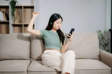 Genç Asyalı kadın evde dinleniyor, kulaklıkla müzik dinliyor ve akıllı telefon kullanıyor.