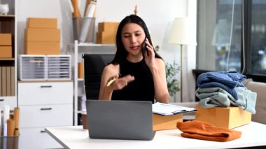  Moda tasarımcısının masasındaki Asyalı genç kadın stüdyoda çalışıyor, dizüstü bilgisayar kullanıyor ve akıllı telefondan konuşuyor. 