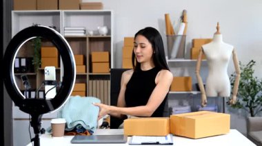Küçük işletme girişimcisi KOBİ serbest. Ev ofisinde çalışan Asyalı bir kadın sosyal medya için paketleme ve akıllı telefon ve yüzük lambası kullanıyor.