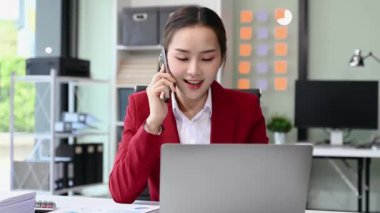Ofiste çalışan Asyalı iş kadını akıllı telefonla konuşuyor ve dizüstü bilgisayar kullanıyor. 