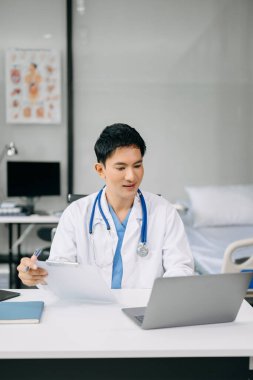 Asyalı Doktor ofis hastanesinde masada oturuyor. İş için dizüstü bilgisayar kullanan erkek terapist 