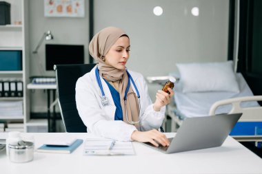 Ofis hastanesindeki masada oturan güzel doktor. Çekici kadın terapist ilaç şişesini tutuyor ve iş için dizüstü bilgisayar kullanıyor. 