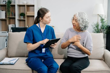 Hemşire ve yaşlı kadın hasta evde dijital tablet kullanıyorlar. Kanepede oturan yaşlı kadınla ilgilenen bir terapist. Tıbbi servis kavramı
