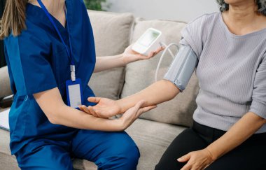 Doktor yaşlı hastayı tansiyon ölçer kullanarak muayene ediyor. Kanepede oturan yaşlı kadınla ilgilenen bir terapist. Tıbbi servis kavramı