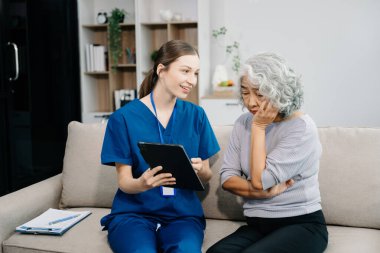 Hemşire ve yaşlı kadın hasta evde dijital tablet kullanıyorlar. Kanepede oturan yaşlı kadınla ilgilenen bir terapist. Tıbbi servis kavramı