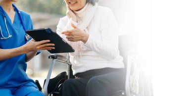 Tekerlekli sandalyedeki yaşlı Asyalı son sınıf öğrencisi. Asyalı dikkatli bir bakıcı ve hastane bahçesinde tablet tableti var.