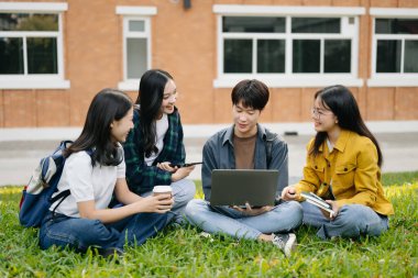 Genç Asyalı üniversite öğrencileri sabah kampüs parkında çimlerin üzerinde otururken ders çalışıyorlar. Birlikte vakit geçirmek, dizüstü bilgisayar ve dijital tablet kullanmak.