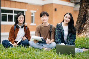 Asyalı öğrenciler kampüs parkında ders çalışıyorlar. Gençler birlikte zaman geçiriyorlar çimenlerde oturuyorlar, kitap okuyorlar, yeşil parkta oturuyorlar.