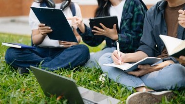 Genç üniversite öğrencileri sabahları çimde otururken kampüs parkında ders çalışıyorlar. Birlikte vakit geçirmek, dizüstü bilgisayar ve dijital tablet kullanmak. 