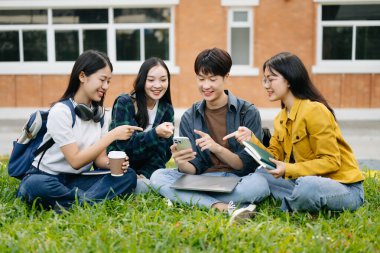 Genç Asyalı üniversite öğrencileri sabah kampüs parkında çimlerin üzerinde otururken ders çalışıyorlar. Birlikte vakit geçirmek, akıllı telefon kullanmak.