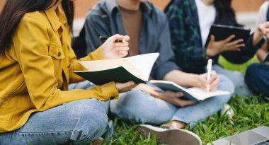 Genç üniversite öğrencileri sabahları çimde otururken kampüs parkında ders çalışıyorlar. Birlikte vakit geçirmek, dizüstü bilgisayar ve dijital tablet kullanmak. 