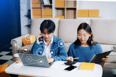 E-ticaret simgeleri, küçük işletmelerin iki kadın sahibinin online siparişleri kontrol etmek, ürünleri satmak ve evdeki kanepede karton kutularla çalışmak için dizüstü bilgisayar kullanması üzerine.