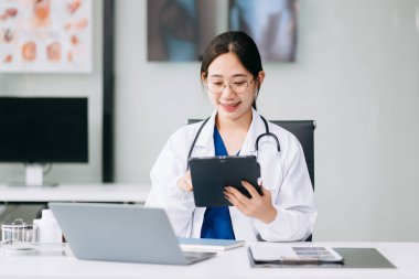 Beyaz üniformalı, kendine güvenen Asyalı kadın doktor masa başında bilgisayar üzerinde çalışıyor. Klinikteki tıp dergisine gülümseyen dizüstü bilgisayar yazıları.