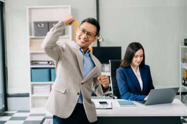 Genç tezahürat yapan Asyalı iş adamı kameraya bakarken iş kadını masada oturuyor ve modern ofiste dizüstü bilgisayarla çalışıyor.