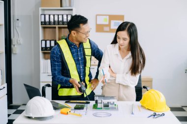 Asyalı mühendisler miğferler, planlar, ev modelleri ve dizüstü bilgisayarla birlikte ofiste mimari projeyi tartışıyor ve birlikte çalışıyorlar. Modern ofiste Asya endüstrisi profesyonel takımı.
