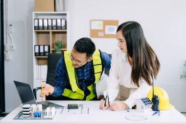 Asyalı mühendisler miğferler, planlar, ev modelleri ve dizüstü bilgisayarla birlikte ofis masasında mimari projeler üzerinde tartışıyor ve birlikte çalışıyorlar.. 