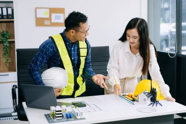 Asyalı mühendisler miğferler, planlar, ev modelleri ve dizüstü bilgisayarla birlikte ofiste mimari projeyi tartışıyor ve birlikte çalışıyorlar. Modern ofiste Asya endüstrisi profesyonel takımı.