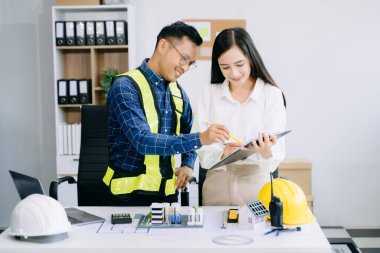 Asyalı mühendisler miğferler, planlar, ev modelleri ve dizüstü bilgisayarla birlikte ofiste mimari projeyi tartışıyor ve birlikte çalışıyorlar. Modern ofiste Asya endüstrisi profesyonel takımı.