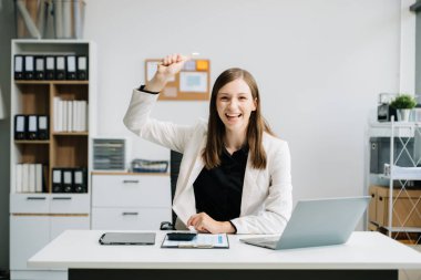 Genç, güzel bir kadın iş yerinde otururken dizüstü bilgisayar kullanarak el kol hareketi yapıyor. Modern ofiste işe odaklanmış.