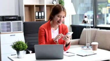 Asyalı iş kadını akıllı telefon kullanıyor ve ofiste dizüstü bilgisayarla çalışıyor. 