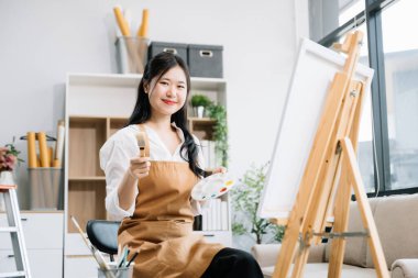 Asyalı kadın ressam sanat atölyesinde resim yapıyor, boya malzemeleri, yağ boyaları kullanıyor ve sanat stüdyosundaki kamera yaratıcı alanında poz veriyor. 