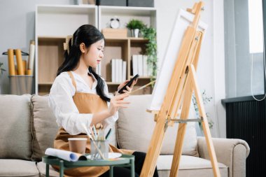 Sanat atölyesinde resim yapan Asyalı kadın ressam, akıllı telefon ve boya malzemeleri, yağ boyama malzemeleri, sanat stüdyosunda yaratıcı alan kullanıyor. 