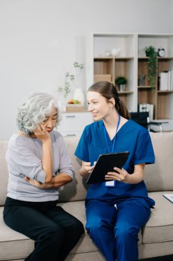 Hemşire ve yaşlı kadın hasta evde dijital tablet kullanıyorlar. Kanepede oturan yaşlı kadınla ilgilenen bir terapist. Tıbbi servis kavramı