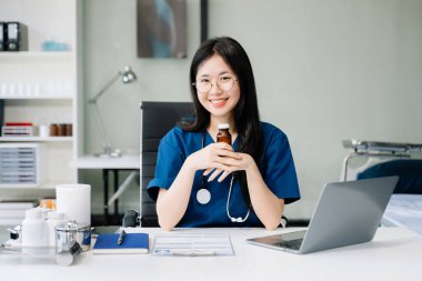 Beyaz üniformalı kendine güvenen genç Asyalı kadın doktor masada oturup dizüstü bilgisayarla ve haplarla çalışıyor.. 