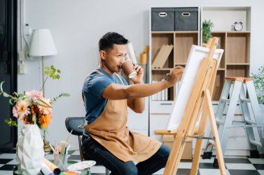 Asyalı erkek ressam sanat atölyesinde resim yapıyor ve kahve içiyor, boya malzemeleri, yağ rengi, tuval sehpası, sanat stüdyosunda yaratıcı alan.