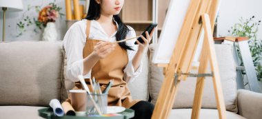 Sanat atölyesinde resim yapan Asyalı kadın ressam, akıllı telefon ve boya malzemeleri, yağ boyama malzemeleri, sanat stüdyosunda yaratıcı alan kullanıyor. 