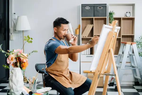 Asian male painter doing artwork in art workshop and drinking coffee, painting supplies, oil pastels, canvas easel, creative space in art studio