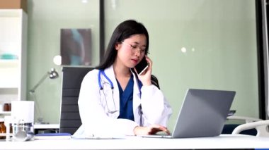 Kendine güvenen genç Asyalı kadın doktor masada oturuyor telefonla konuşuyor ve dizüstü bilgisayar kullanıyor..