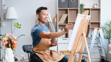 Asyalı erkek ressam sanat atölyesinde resim yapıyor ve kahve içiyor, boya malzemeleri, yağ rengi, tuval sehpası, sanat stüdyosunda yaratıcı alan.