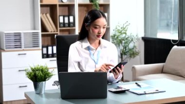 Ofiste çalışan Asyalı iş kadını akıllı telefon ve dizüstü bilgisayar kullanıyor. 