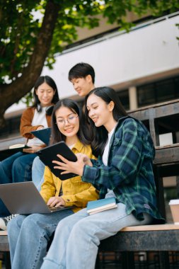 Genç Asyalı üniversite öğrencileri sabahleyin kampüs parkında ders çalışıyor. Birlikte vakit geçirmek, dizüstü bilgisayar ve dijital tablet kullanmak.