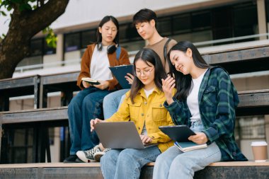 Genç Asyalı üniversite öğrencileri sabahleyin kampüs parkında ders çalışıyor. Birlikte vakit geçirmek, dizüstü bilgisayar ve dijital tablet kullanmak.