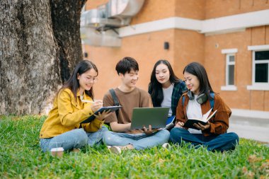 Genç Asyalı üniversite öğrencileri sabah kampüs parkında çimlerin üzerinde otururken ders çalışıyorlar. Birlikte vakit geçirmek, dizüstü bilgisayar ve dijital tablet kullanmak.