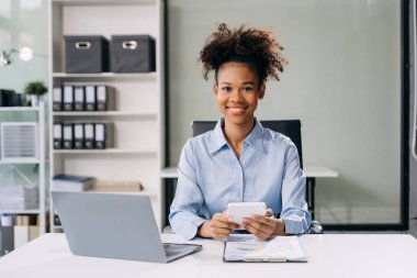 Gülümseyen, hesap makinesi olan ve modern ofiste dizüstü bilgisayar kullanan kendine güvenen bir iş kadını.