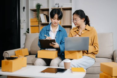 Küçük işletmelerin iki kadın sahibi online siparişleri kontrol etmek için dizüstü bilgisayar ve tablet kullanıyorlar, ürünler satıyorlar, kanepede karton kutularla çalışıyorlar.