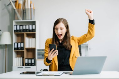 Kendine güvenen bir iş kadını, yaratıcı ofiste masa başında otururken akıllı telefon kullanır ve el kol hareketi yapar. 