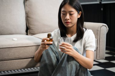 Depresyondaki Asyalı kadın koltukta oturmuş elinde bir şişe hap ve su bardağı tutuyordu.