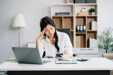 Asyalı iş kadını, modern ofiste akıllı telefon ve dizüstü bilgisayarla çalışmaktan stresli, sıkılmış ve aşırı düşünüyor.