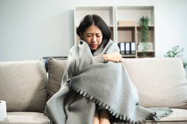 Kanepede battaniyeyle kaplanmış, grip belirtileri gösteren genç bir Asyalı kadın. Hastalık, sağlık ve iyileşme kavramı