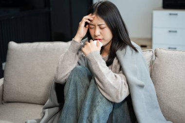 Kanepede battaniyeyle kaplanmış, grip belirtileri gösteren genç bir Asyalı kadın. Hastalık, sağlık ve iyileşme kavramı